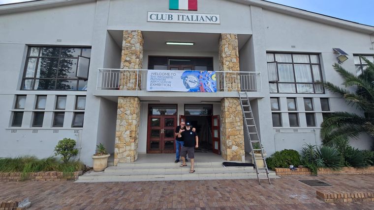 Two people on the steps of a venue beneath a banner welcoming visitors to the Inclusive Plumbers Association exhibition